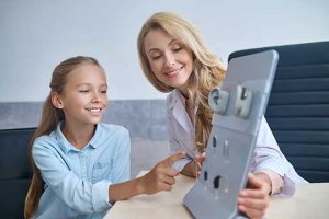 A smiling woman shows a young girl different hearing aid models on a display board in an office setting, likely after a recent hearing test. The girl points at the board while both look happy and engaged.